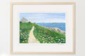 Watercolor of seaside cottage with path along the ocean, winding through grass and wildflowers, shown in a wood frame.
