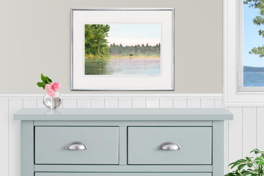 Watercolor painting of a moose at the edge of a pond with trees. Shown over a gray dresser.