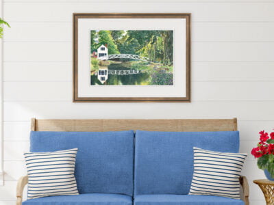 White arched footbridge in Somesville Maine reflecting in a pond with trees and flowers. Shown framed over a blue bench.
